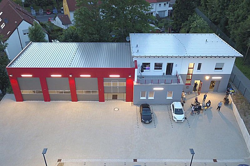Luftbild des Gerätehauses in der Dämmerung: rote Fahrzeughalle links, weißes Sozialgebäude rechts, großer Übungshof davor.